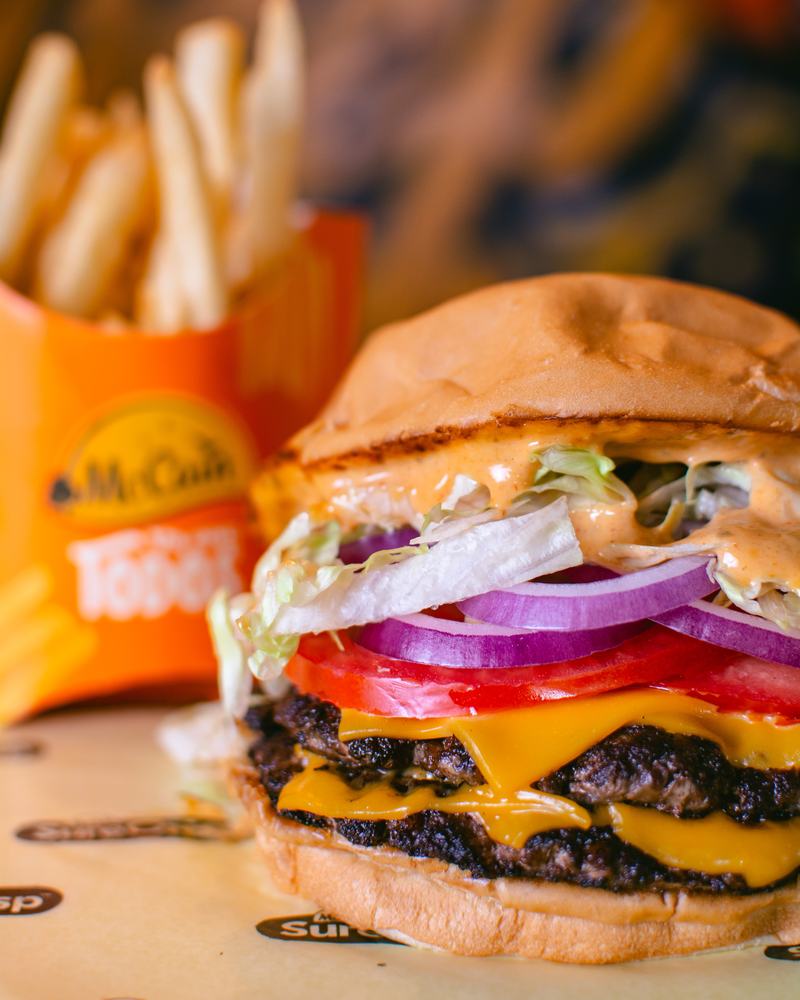Hamburguesa doble con queso cheddar, cebolla morada y tomate, con papas fritas de fondo, producto de Burger Cheep
