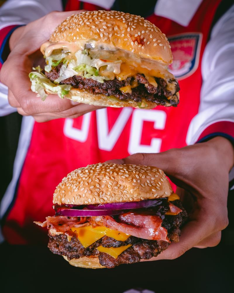 Dos hamburguesas de Burger Cheep sostenidas con las manos, una con lechuga y salsa y otra con panceta y cebolla morada